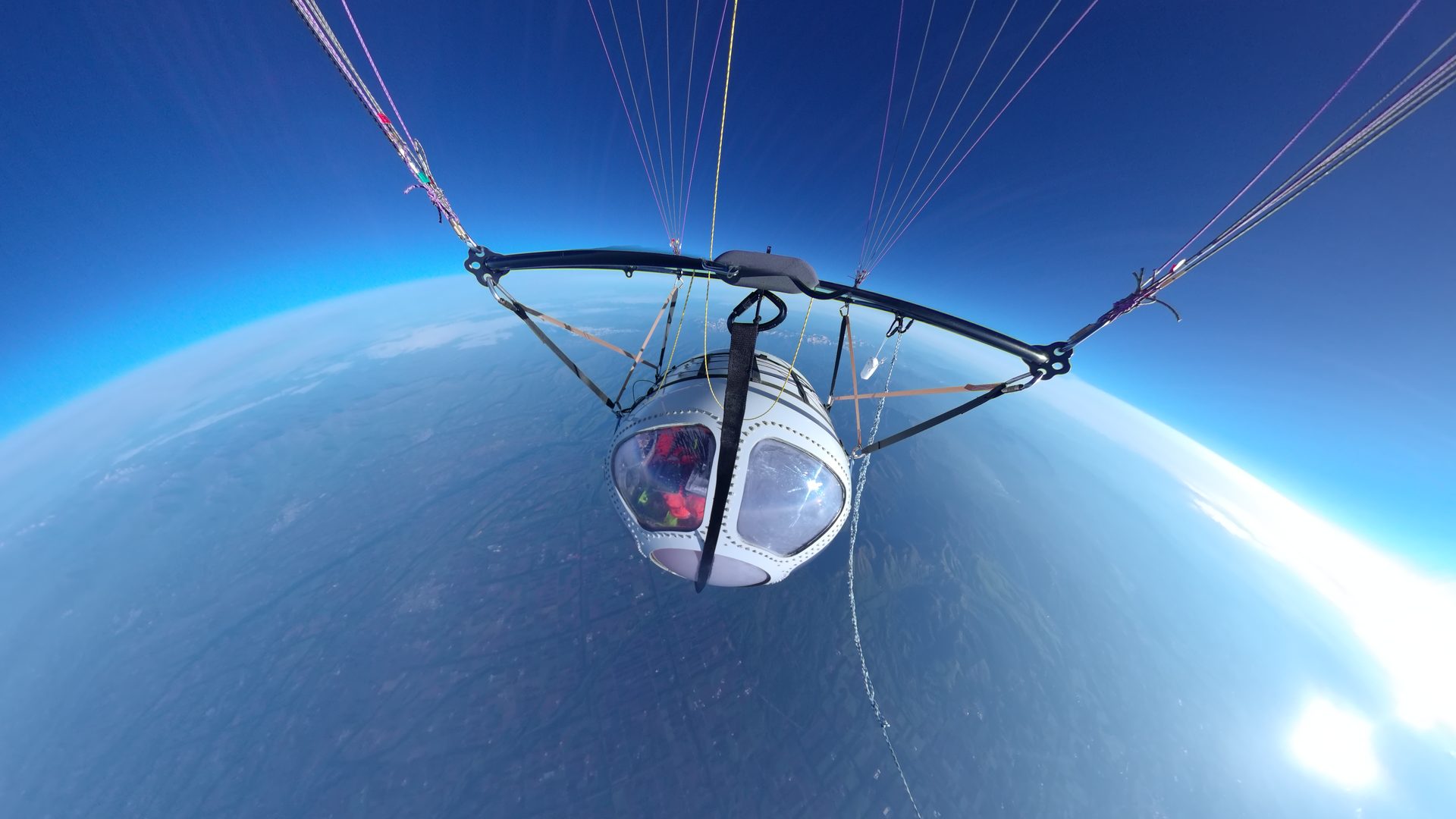 View from a paraglider cockpit high above Earth, with colorful suspension lines extending to the canopy and a curved blue horizon below.