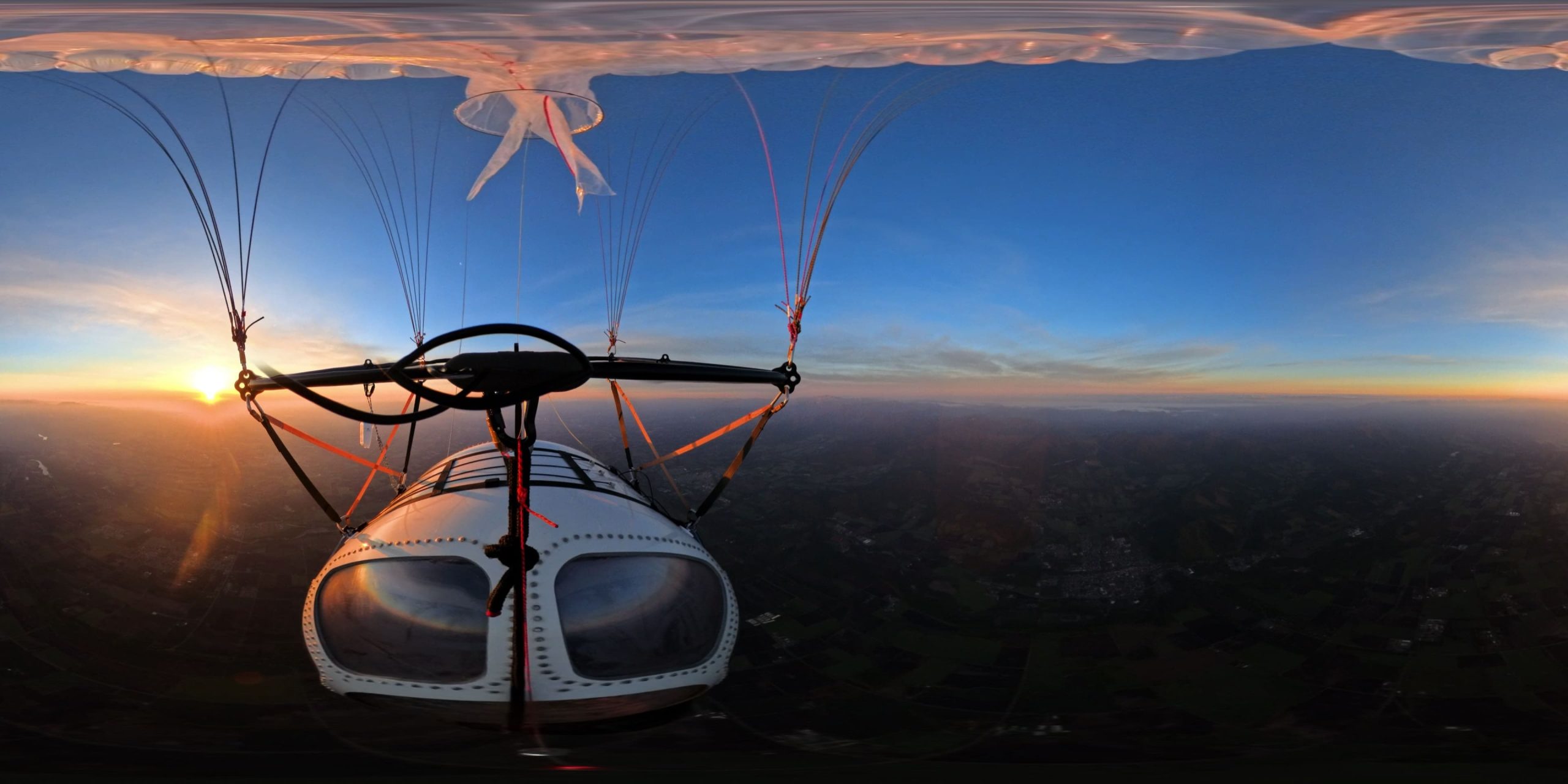 Front view from a small paraglider cockpit during sunset, with lines overhead and a sunlit horizon below over farmland loinography,