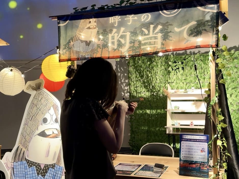 Person in silhouette aiming a rifle at an indoor exhibit booth with a Japanese banner and decorative greenery.