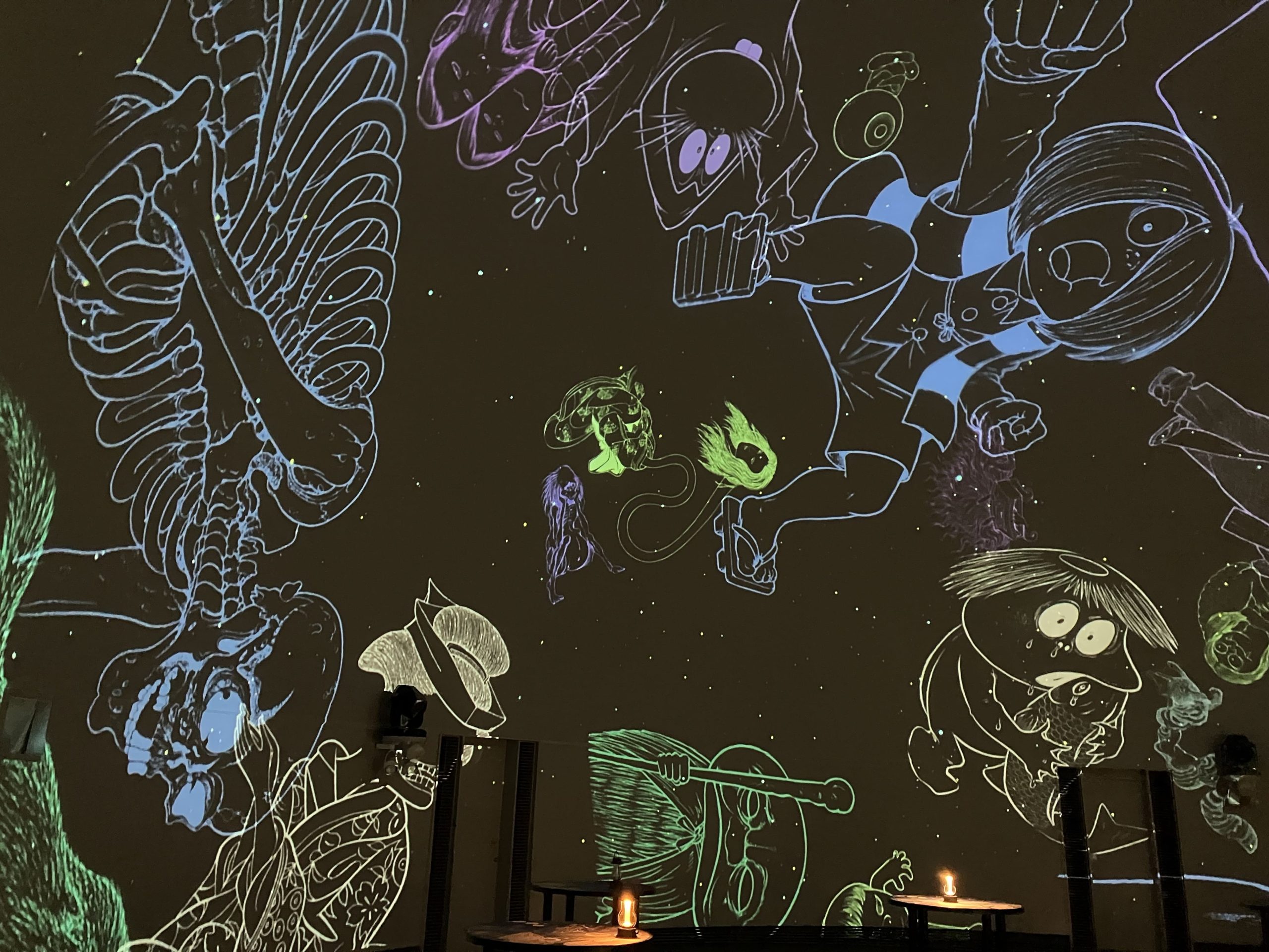Dimly lit cafe with a dark wall covered in glowing chalk outlines of cartoon characters and round candlelit tables nearby.