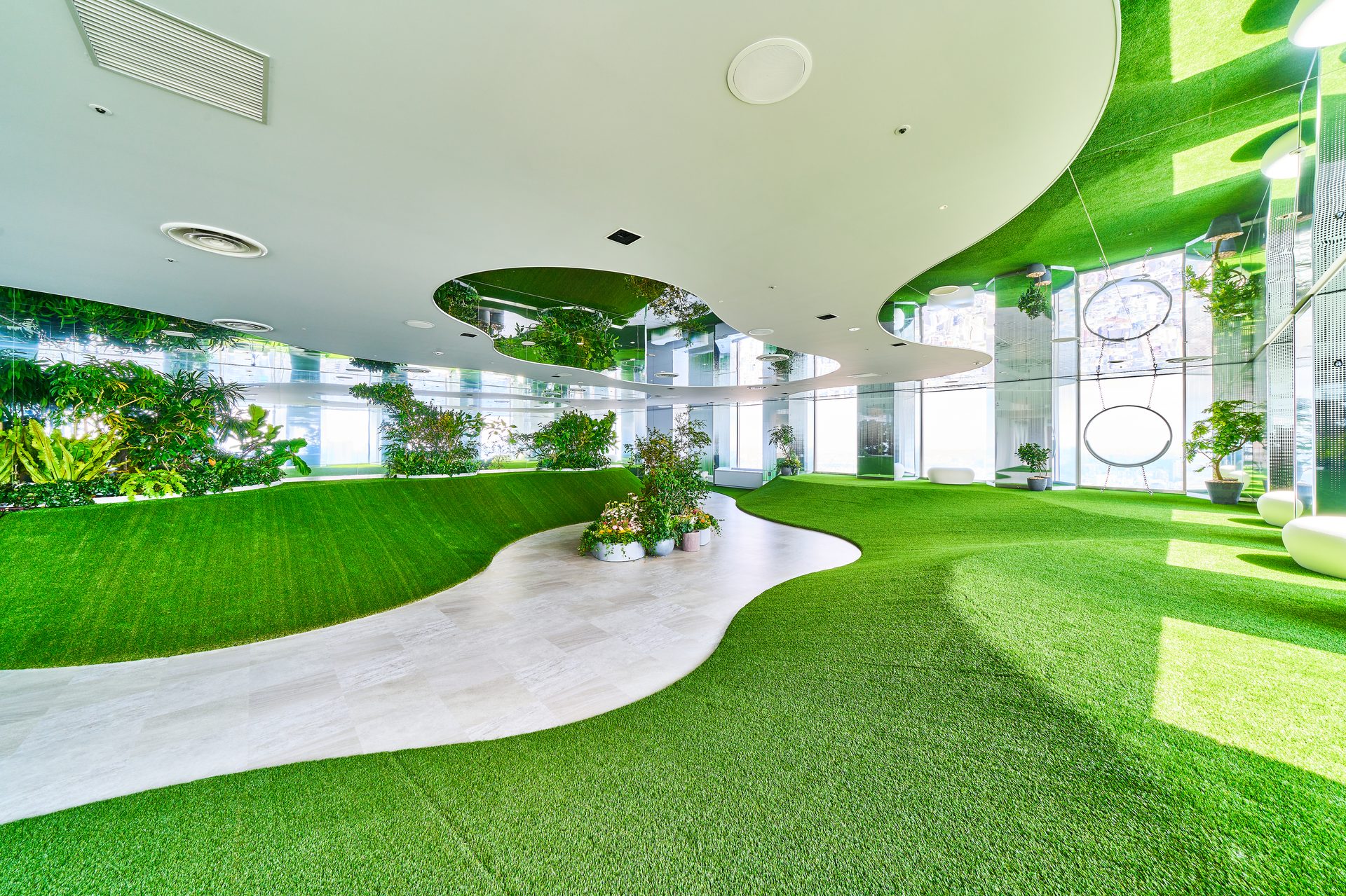 Indoor green garden lobby with artificial grass, curved white path, and large windows letting in daylight.