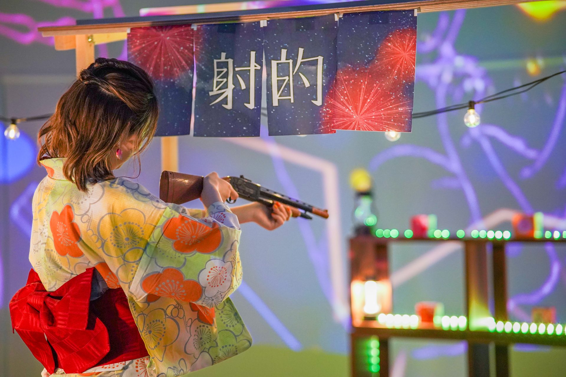 Person in a bright floral kimono aiming a prop rifle at a festival, banners and colorful lights in the background.
