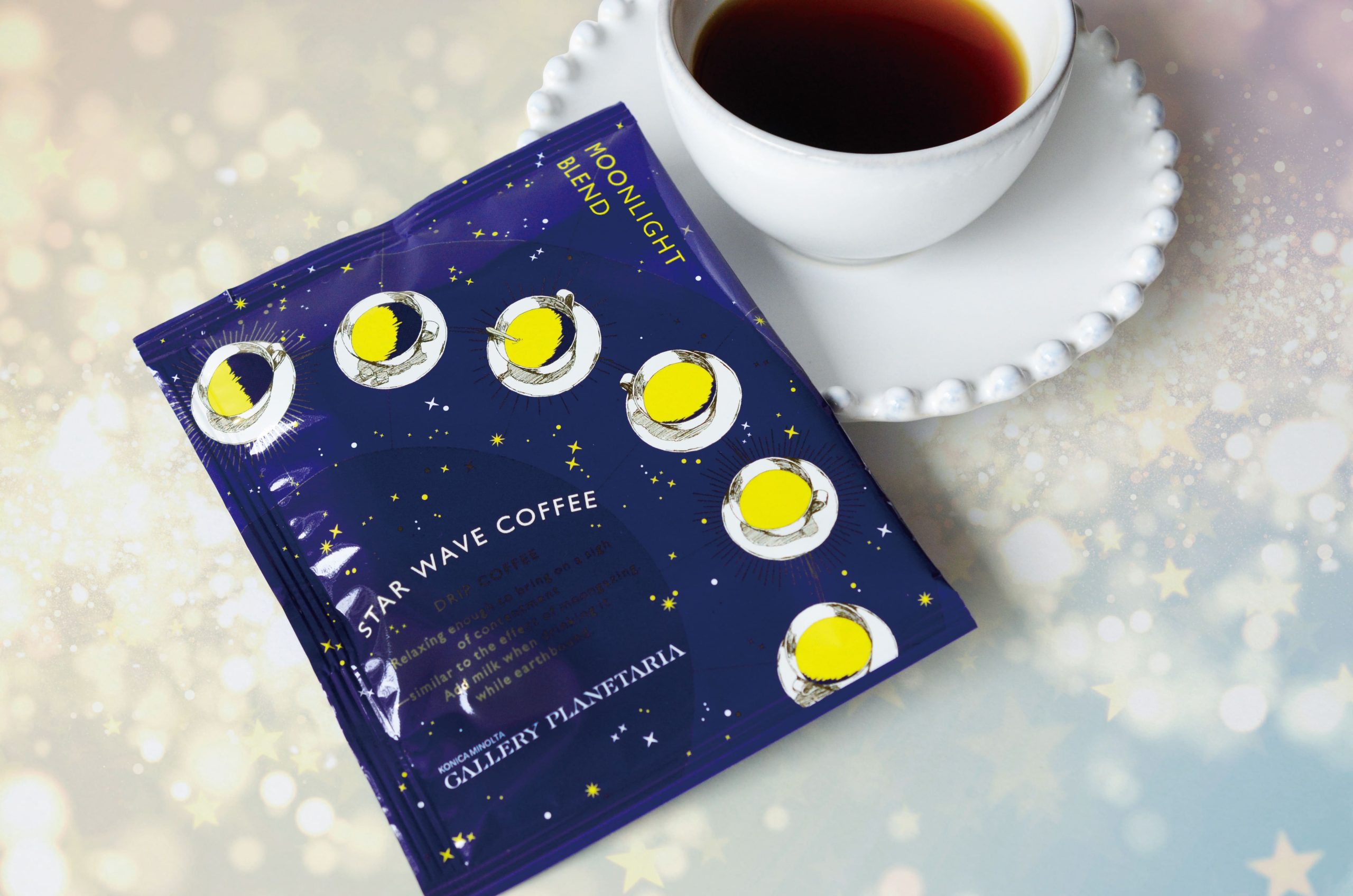 Blue Star Wave Coffee package with yellow moon illustrations beside a white cup of black coffee on a decorative white saucer, diagonal view.