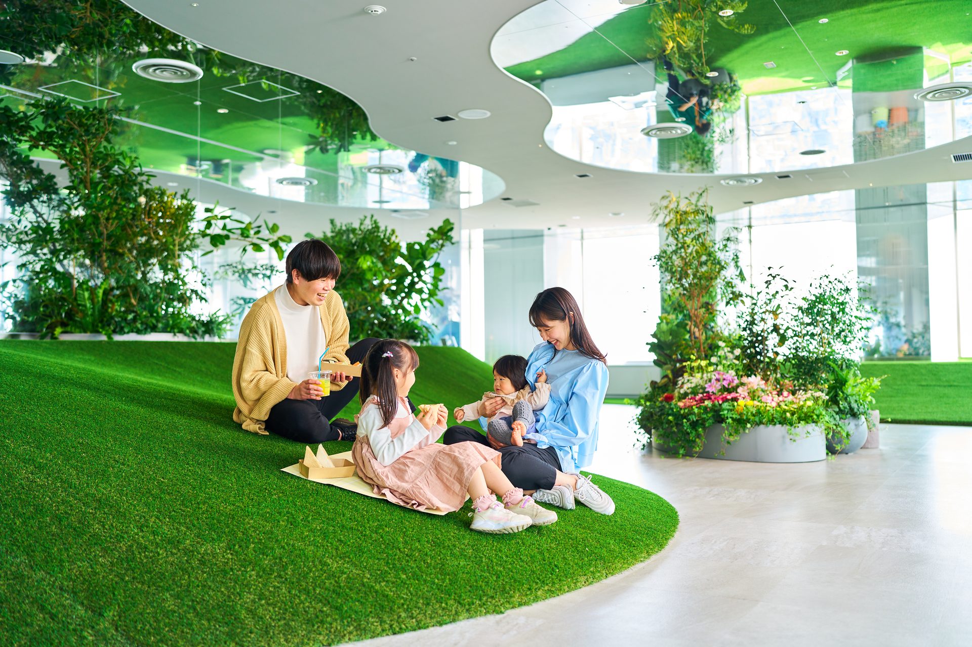 Family sitting on indoor artificial grass, sharing snacks with two adults and two children amid lush plants.