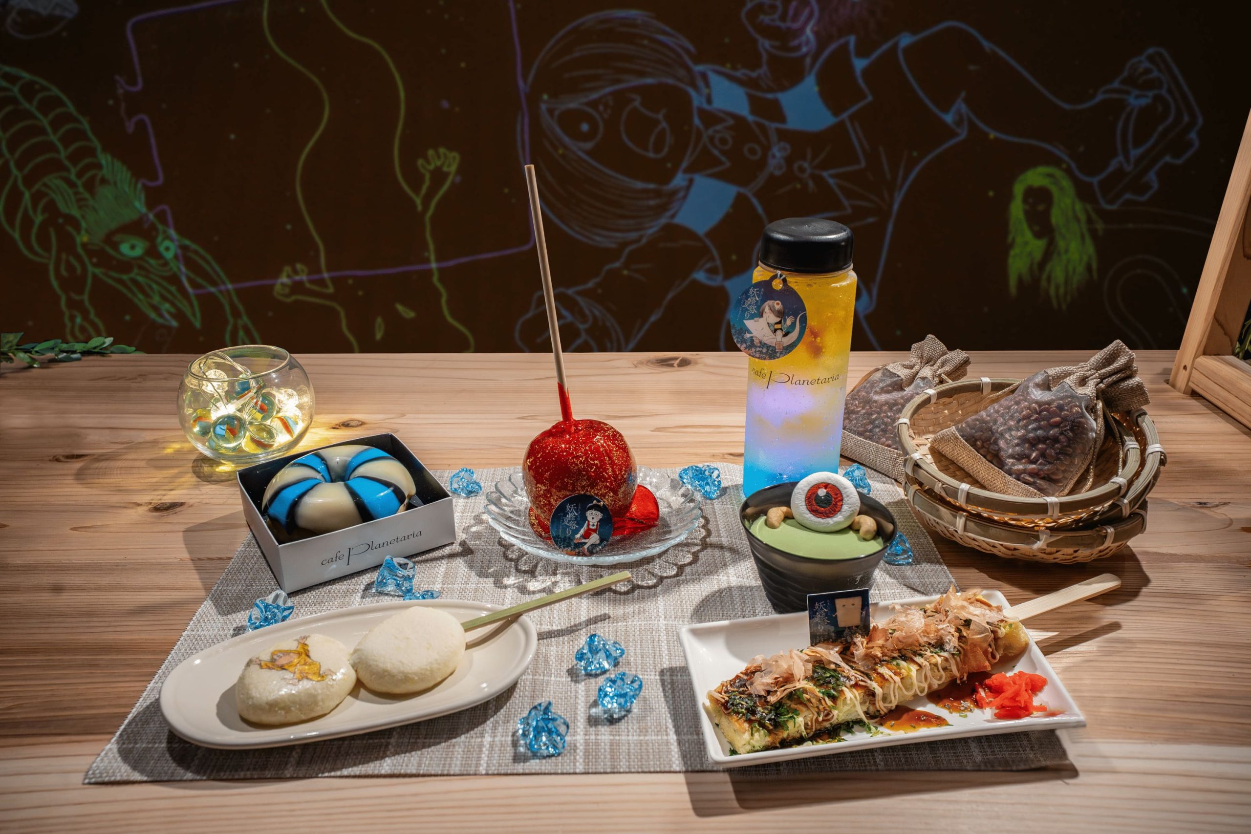 Dessert table on a wooden surface with a red candied apple on a stick, blue-green candies, a blue-and-white swirled pastry in a box, and a bottle labeled Café Planetaria.