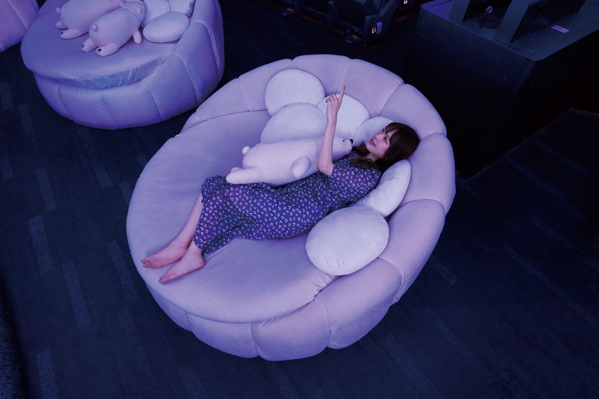 Woman relaxing on a large lavender bean bag, hugging a big plush unicorn pillow in a dimly lit room.